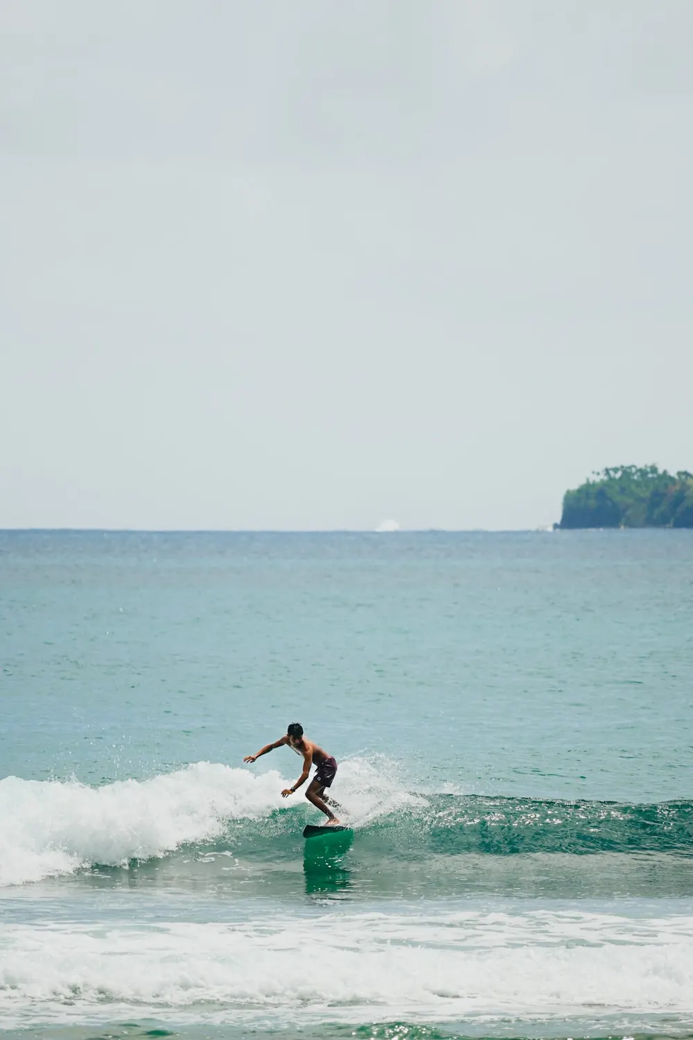A surfer riding a small green wave with arms out for balance and an island on the horizon